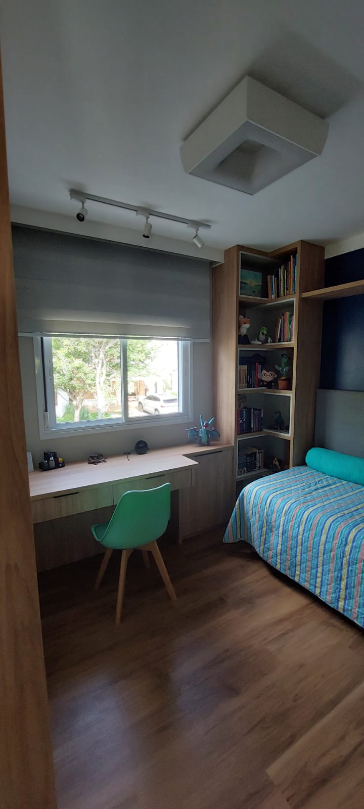 Quarto juvenil com escrivaninha sob janela, estante de livros e cama listrada