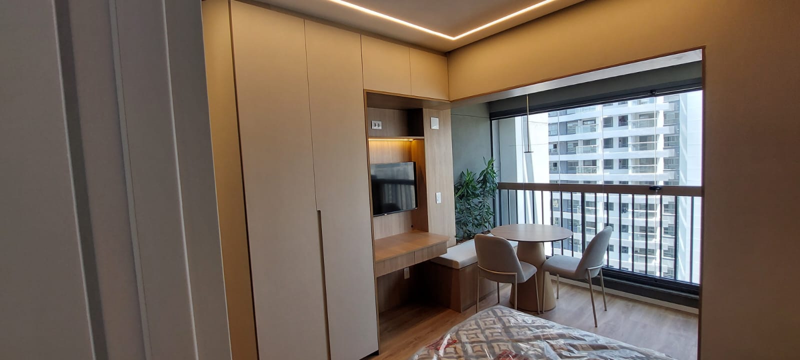 Studio bege com painel de TV, bancada e mesa na varanda