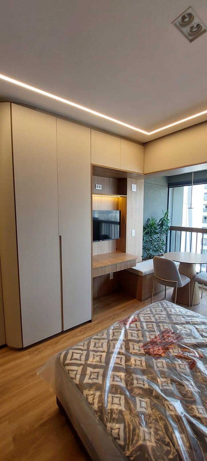 Studio com armários bege, painel de TV e sanca de LED; colchão ainda plastificado